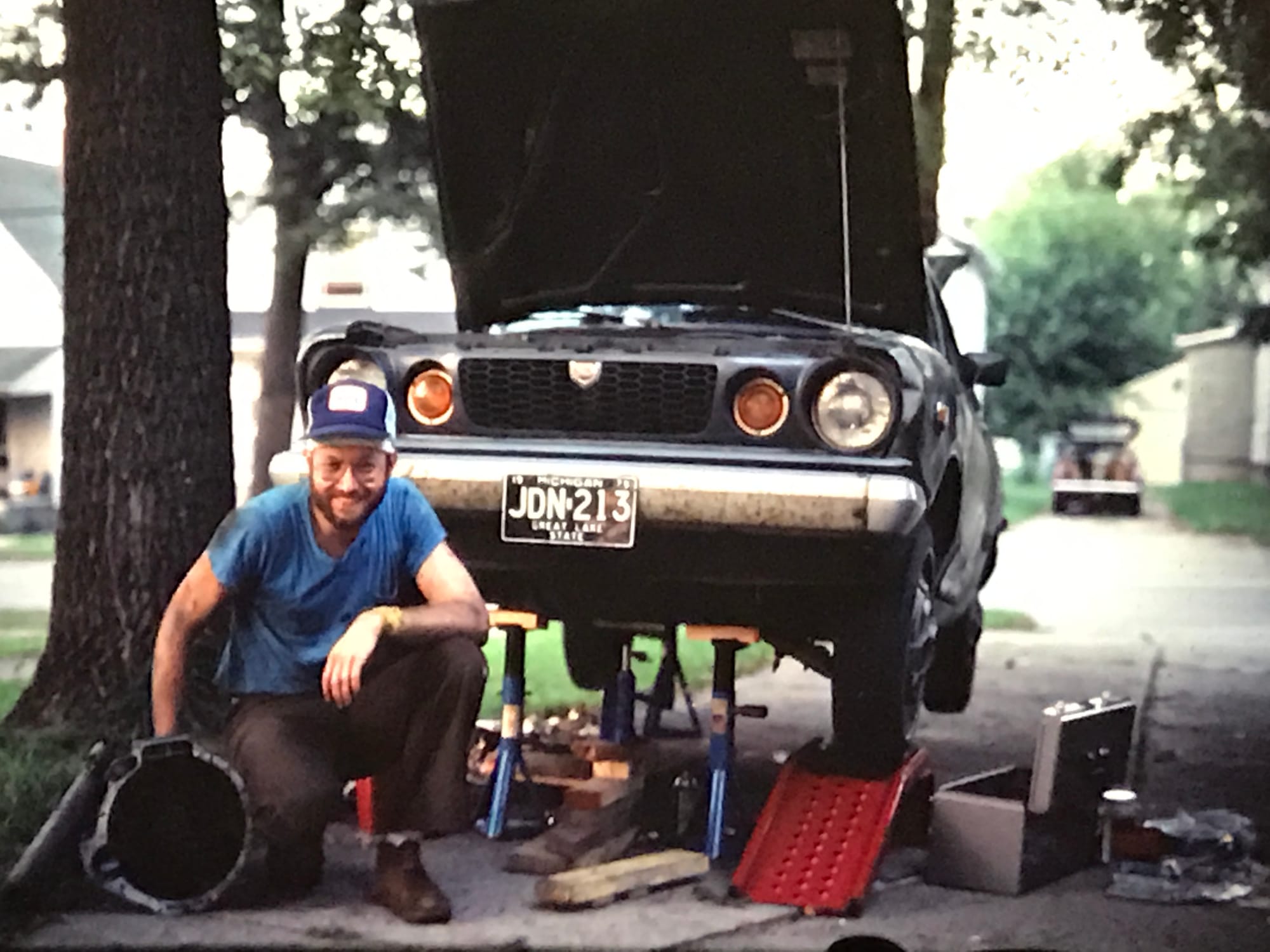 White man wearing a blue trucker cap, stained shirt, brown slacks. He crouches by a gray car on jacks, tools scattered nearby