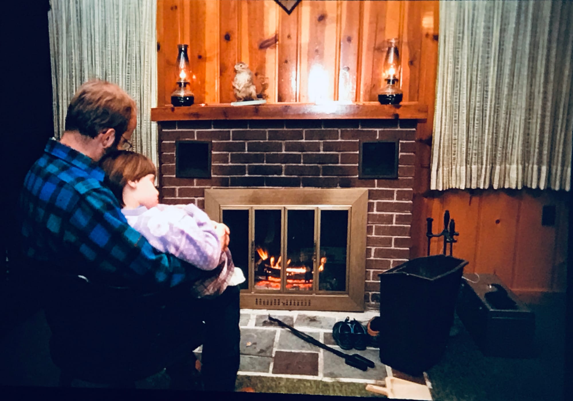 A blond bearded man in a blue flannel holds me (brown hair, purple sweater) on his lap in front of a brick fireplace.