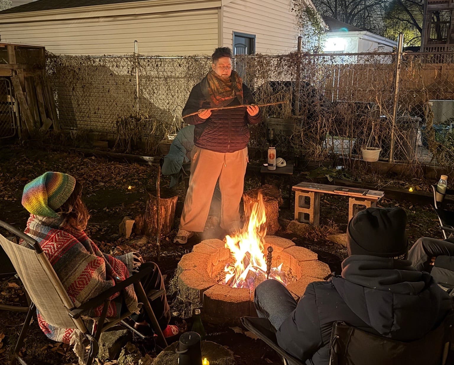 Julia stands by the fire pit, eyes closed holding a stick into which she sends her love and gratitude. People sit around the fire facing Julia.