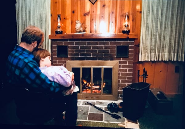 A blond bearded man in a blue flannel holds me (brown hair, purple sweater) on his lap in front of a brick fireplace.